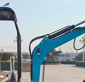 A close-up detail of a turquoise mini excavator boom, showing black hydraulic cylinders, chrome bolts, protective hose sleeves, and the adjacent operator control joystick under an open canopy.