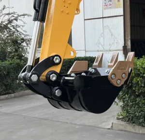 Close-up detail of a heavy-duty black digging bucket with replaceable teeth, connected to a yellow mini excavator arm via a hydraulic quick coupler.