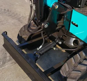 Close-up detail of a black heavy-duty dozer blade with hydraulic cylinder and protective hose sleeves on a turquoise mini excavator.