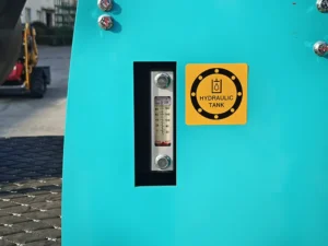 A close-up of a clear hydraulic fluid level gauge with numerical markings on a turquoise mini excavator, next to a yellow ‘Hydraulic Tank’ safety label.
