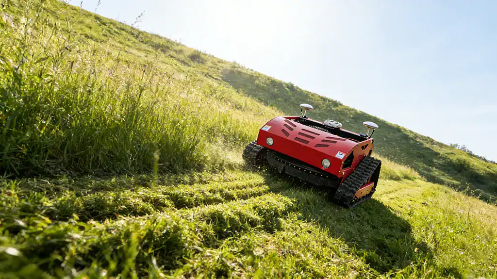 Red remote-controlled tracked lawn mower actively cutting grass on a steep, sunlit hillside, with visible clippings being discharged.