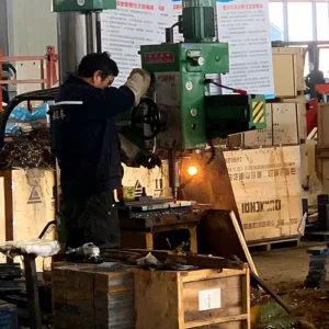 Side view of a worker operating a green vertical drilling machine to process metal components in a factory workshop.