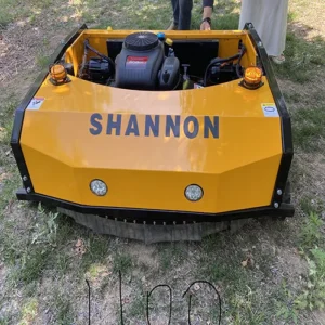 Front side view of a yellow "SHANNON" remote-controlled tracked lawn mower with a 608cc gasoline engine, LED headlights, and warning lights, operating in a grassy woodland area.