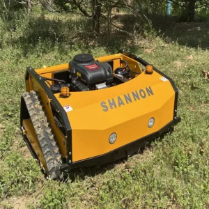 Front side view of a yellow "SHANNON" remote-controlled tracked lawn mower with LED headlights and warning lights, operating in a grassy woodland area.