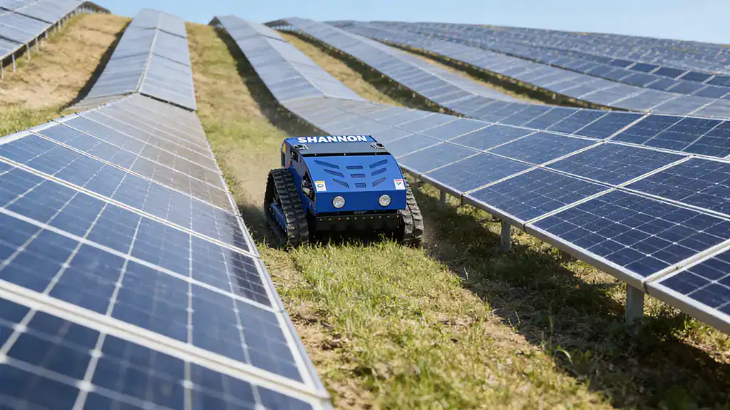 Blue SHANNON remote-controlled tracked lawn mower operating in a sloped solar farm between solar panel arrays.